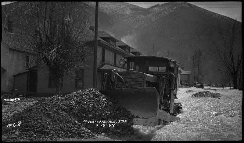 Image of a tractor during a flood in Wallace, Idaho. It appears to be have been used to move rocks. Some standing buildings are in the background.