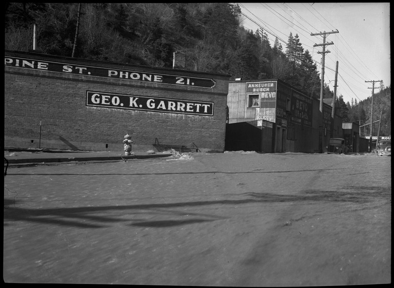Image of several buildings. The closest building reads " Pine St. Phone 21." and "Geo. K. Garrett." Water covers all of the street. Two vehicles can be seen on the right. 