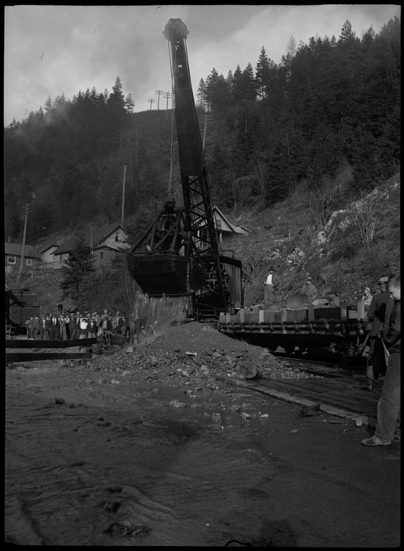 A crowd of people standing behind a fence watching a large crane, possibly during a flood. There is some flowing water approaching the pile of material. There are a few cartloads of more material (rocks, debris) on a conveyor belt on the right.