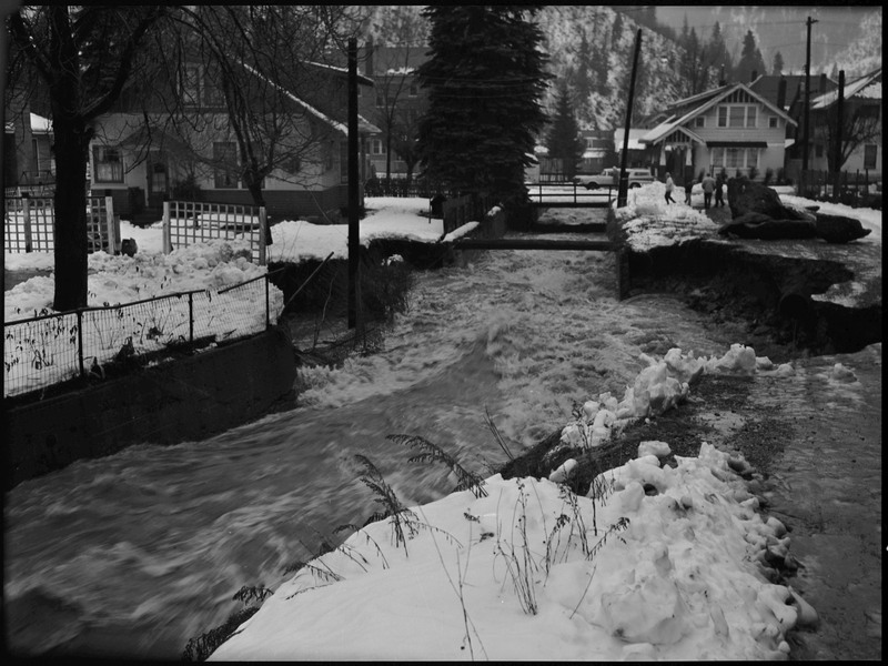 Water rushing through Wallace, Idaho during a flood. A few people are walking in the distance on the right. There is snow on the ground and on surrounding buildings.