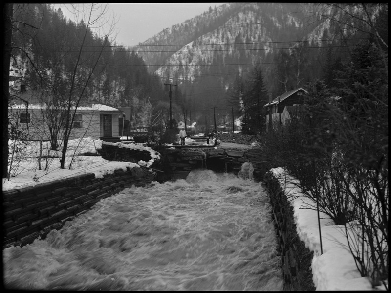 Water rushing during a flood in Wallace, Idaho. The water is currently contained by stacked lumber. Some buildings are still standing in the distance. Snow covers the ground and the buildings. 