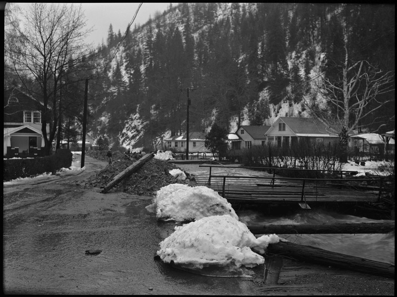 Street view of a bridge and surrounding buildings during a flood. There are piles of snow along the bank of the flowing water. Piles of material (rock, debris) line the bank to prevent flooding into the buildings.