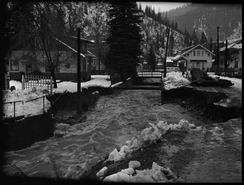 Water rushing through Wallace, Idaho during a flood. Water has eroded parts of the ground. A few people are walking in the distance on the right. There is snow on the ground and on surrounding buildings.