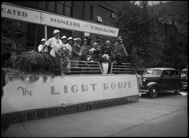 People on the Lighthouse parade float. Several people wear hardhats and others are in suits. Text on the float reads, " [Dedi]cated to the Pioneer of the Coeur D'alenes The Lighthouse."