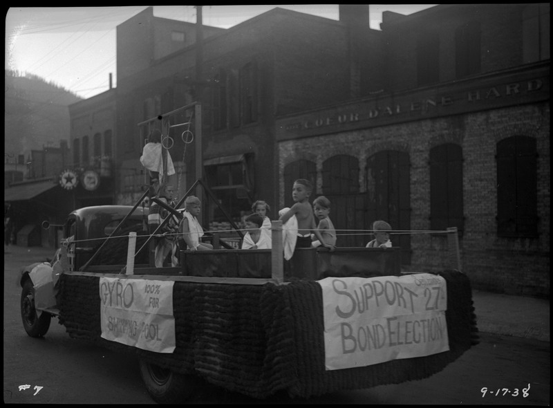 Children sitting on the Wallace Gyro club parade float during the Benevolent and Protective Order of Elks Roundup parade. They are wearing towels around their bodies. A set of what looks like playground rings are on the float. Text of the float reads "Gyro 100% for Swimming Pool" and "Support [illegible] 27th Bond Election."