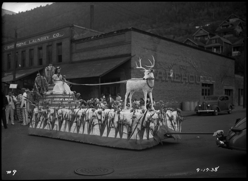 A girl sits on a chair appearing to pull a fake elk on the Elks float during the BPOE (Benevolent Protective Order of the Elks) Roundup parade. The girl is wearing a white dress and the float is adorned with flowers. The Shoshone Laundry Company building is in the background.