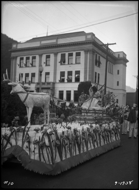A girl sits on a chair appearing to pull a fake elk on the Elks float during the BPOE (Benevolent Protective Order of the Elks) Roundup parade. The girl is wearing a white dress and the float is adorned with flowers. The girl is turning away from the camera.