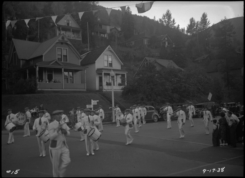 The Elks band performing during the Benevolent and Protective Order of Elks Roundup parade. They are wearing identical white suits. Houses on a hill are in the background. Some people can be seen watching the band perform from a porch.