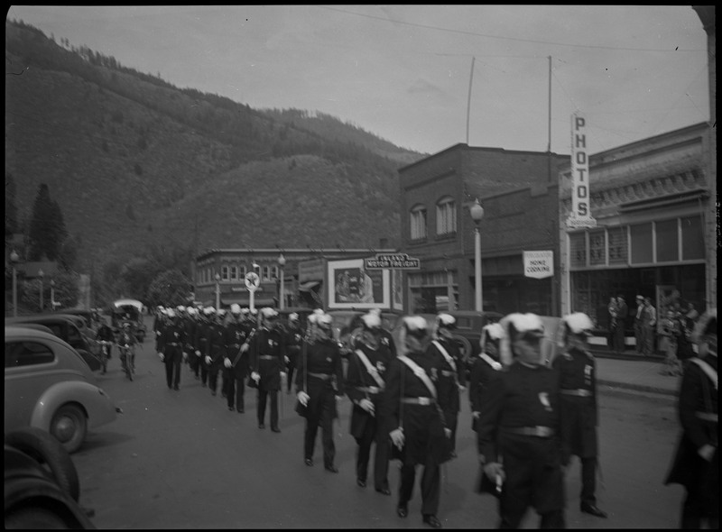 Men in uniform marching in Benevolent and Protective Order of Elks parade. Cars are parked along the sides of the street.
