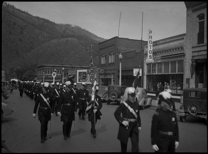 Men in uniform marching in Benevolent and Protective Order of Elks parade as spectators watch on the sidewalk. Cars are parked along the sides of the street.