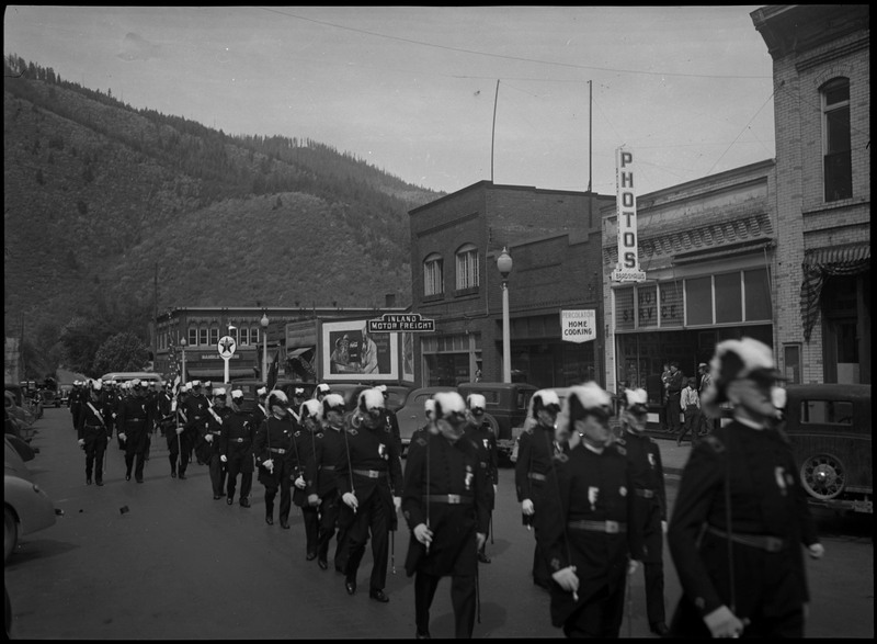 Men in uniform marching in Benevolent and Protective Order of Elks parade as spectators watch on the sidewalk. Cars are parked along the sides of the street.