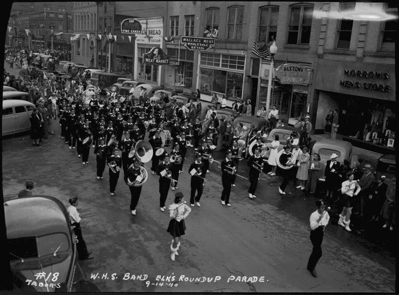 The Wallace High School band performing in the Benevolent and Protective Order of Elks Roundup parade. They are wearing identical uniforms and walking down a street. Spectators watch from the sidewalk. Stores including, Morrows Men's Store and Saxton's Ice Cream and Candy are in the background.