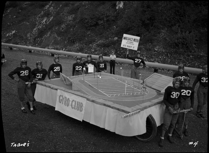 The Gyro club float in the Benevolent and Protective Order of the Elks. Several football players stand in their uniforms around the float. The float is decorated with a mini replica of a football field. One of the players holds a sign that reads, "Wallace Needs An Adequate Athletic Field Let's Boost it." 