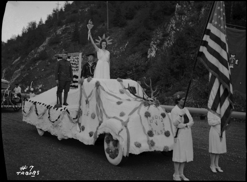 A woman posing as the Statue of Liberty and two uniformed men standing on the Lady liberty float/Aux. V.F.W. float in the Benevolent and Protective Order of the Elks parade. Antlers and a clock decorate the float. Two women holding flags stand in front of the float.