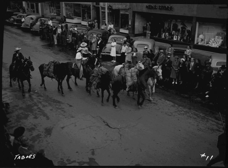 People riding horses down the street in the Elks Roundup parade. Spectators watch by parked automobiles and on the sidewalk.