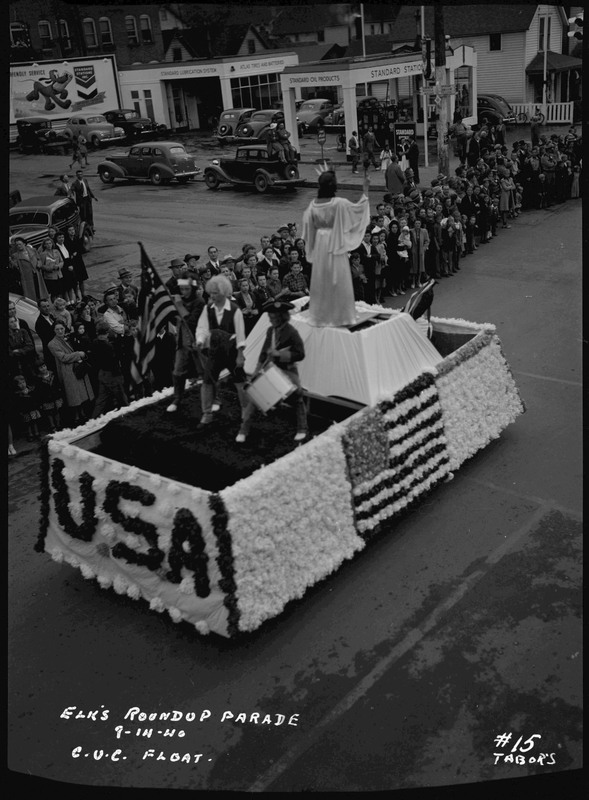 The back of the C.U.C. float in the Elks Roundup parade. A woman, posing as the Statue of Liberty, and three people wearing colonial style dress stand on the float. Spectators watch the float on the side of the street.