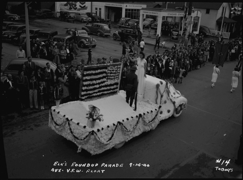 The Aux V.F.W. float in the Elks Roundup parade. A woman wearing a dress and a uniformed man stand on the float which is decorated with an American flag. Spectators watch from the side of the street.