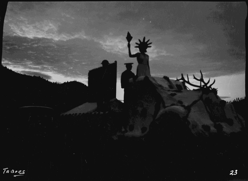A darkened image of the Aux. V.F.W. float in the Elks Roundup parade. A woman dressed like the Statue of Liberty and two other people can be seen as dark silhouettes on the float.