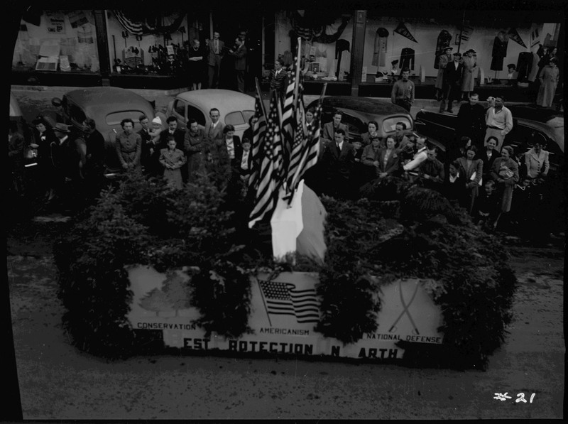 The Forest Service float decorated with foliage and American flags in the Elks Roundup parade as spectators watch near parked automobiles.
