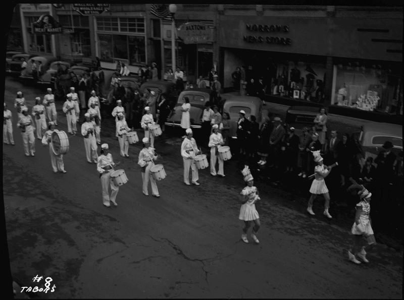 The Wallace High School band in the Elks roundup parade. The band members are wearing identical white uniforms. Spectators watch from the parked automobiles on the side of the street.