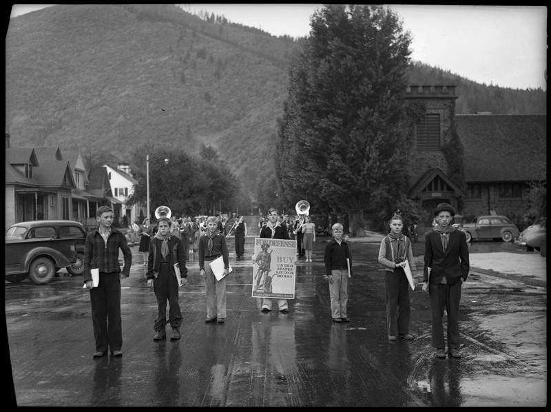 Children standing in the street with war bonds posters in Defense Bond Parade. One of the posters read, "For Defense BUY United States Savings Bonds On Sale At Your Post Office or Bank." Band members stand with their instruments behind the children.