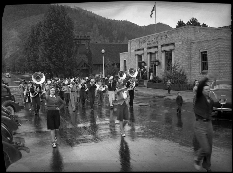 Band playing in Defense Bond Parade. They are passing by the Wallace Post Office, where a group of people are watching the band walk by.