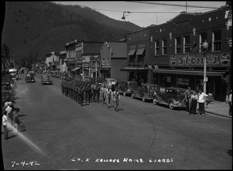 Co. K. Kellogg home guards marching in a July 4th parade. Spectators stand by parked automobiles to watch the parade.