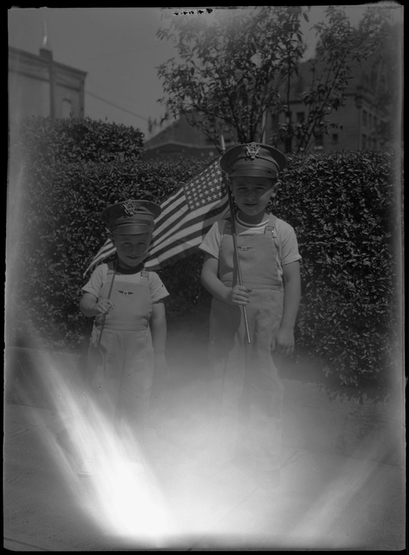 Children wearing matching overalls and hats and holding American flags for 4th of July Parade. The bottom of the image is bright from lens flare.