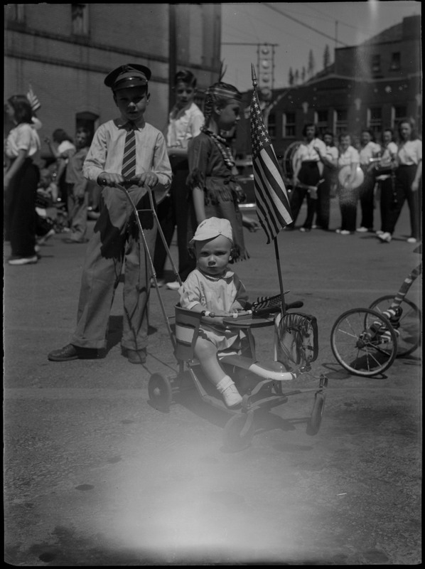 A child dressed in a suit and hat push a toddler sitting in a stroller during a July 4th parade. The toddler is holding an American flag and there is another American flag attached to the stroller. Other children and a group of women holding instruments are in the background.