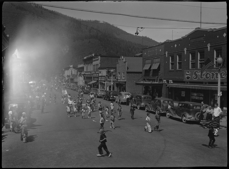 Children in July 4th parade, some are holding American flags. Spectators watch on from the sides of the street.
