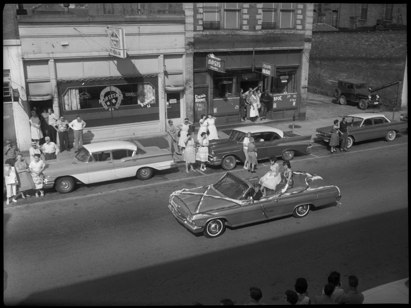 Two women in a car during Benevolent and Protective Order of Elks roundup parade. A man sits in the driver's seat. Spectators watch from the sidewalk.
