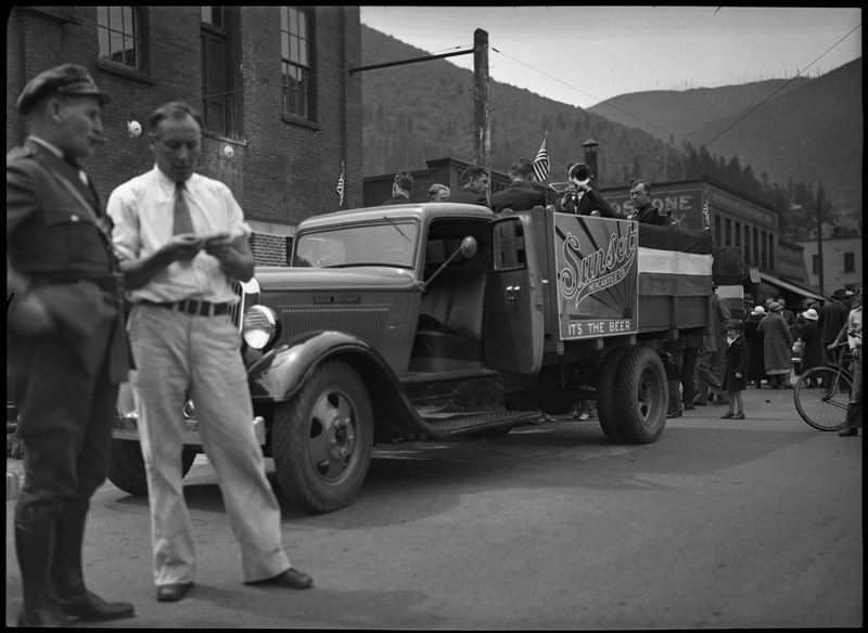 Men sitting in a truck during the Sunset Brewery parade. The side of the truck reads, "Sunset Mercantile Co. It's the Beer." Two men stand close to the camera on the left, one is smoking a cigarette. A crowd of people can be seen in the distance on the right.
