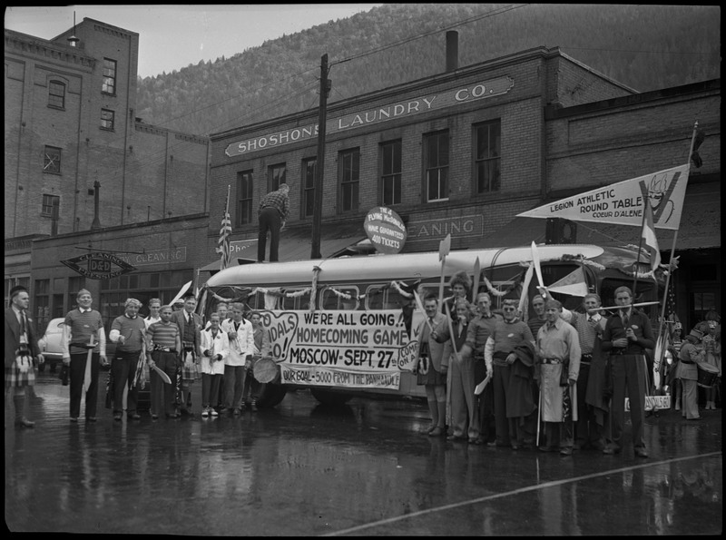 A crowd of people stand around the Legion Athletic Round Table float at the University of Idaho Homecoming Parade. The float is advertising the Homecoming Game, which took place in Moscow on September 27th. The photo was taken outside Shoshone Laundry Co.