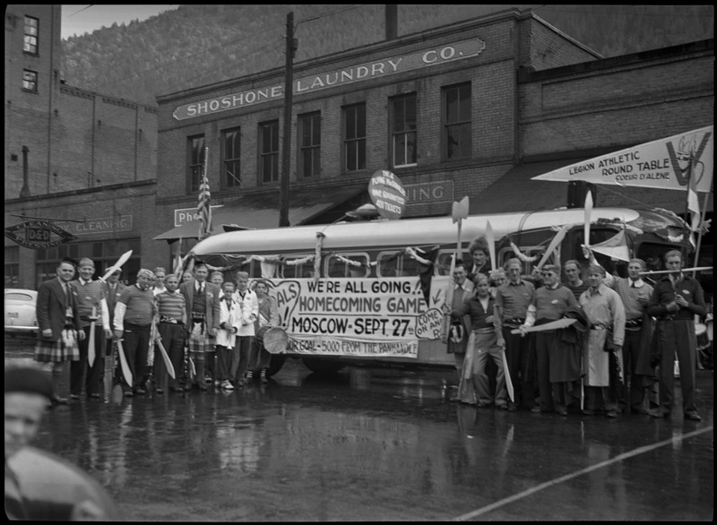 A crowd of people stand around the Legion Athletic Round Table float at the University of Idaho Homecoming Parade. The float is advertising the Homecoming Game, which took place in Moscow on September 27th. The photo was taken outside Shoshone Laundry Co.
