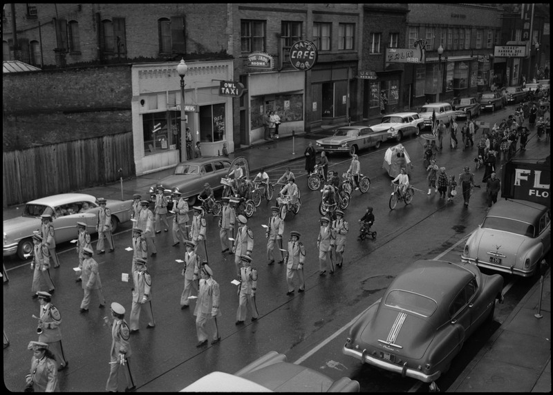 A group of uniformed children walking in the Benevolent and Protective Order of the Elks parade. Some children in costumes are walking behind those in uniform. Cars are parked along the sides of the street.