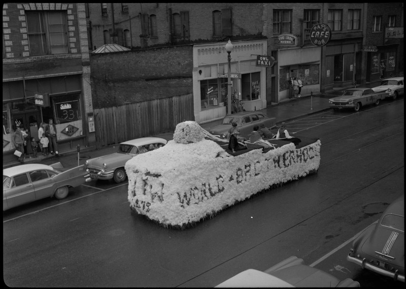 Five individuals sit on a decorated float during the Benevolent and Protective Order of the Elks parade. People can be seen near building watching the procession.