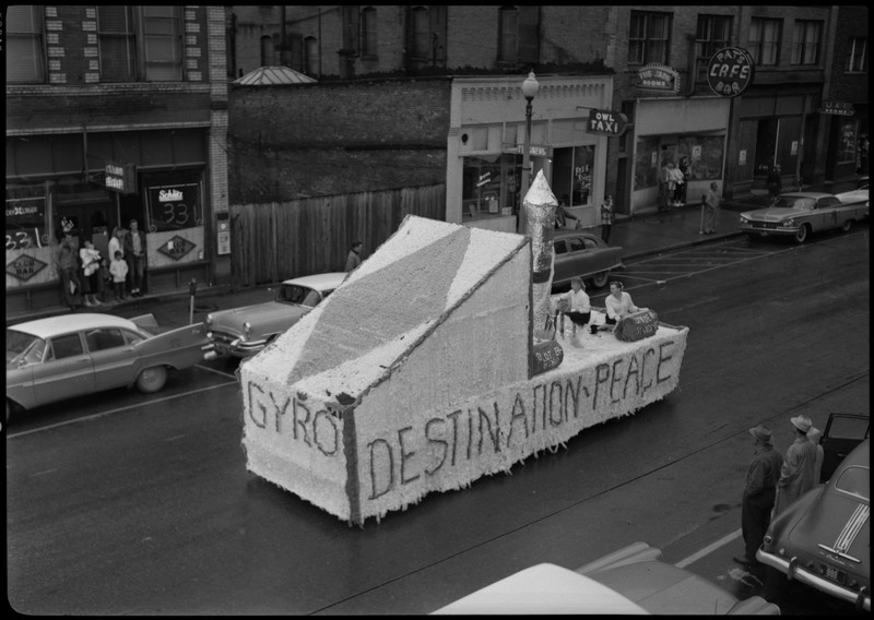 Two girls sit on a Gyro float during the Benevolent and Protective Order of the Elks parade. The side of the float reads, "Destination - Peace." Spectators can be seen on the sides of the street watching the parade.