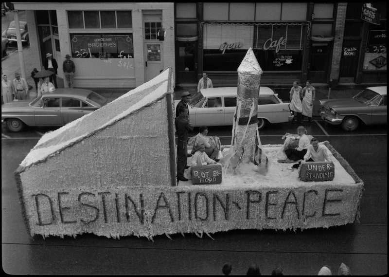 Six individuals sit on a Gyro float during the Benevolent and Protective Order of the Elks parade. The side of the float reads, "Destination - Peace" and two signs on the float read, "Brotherhood" and "Understanding." (The text on the "Brotherhood" sign is misspelled.)