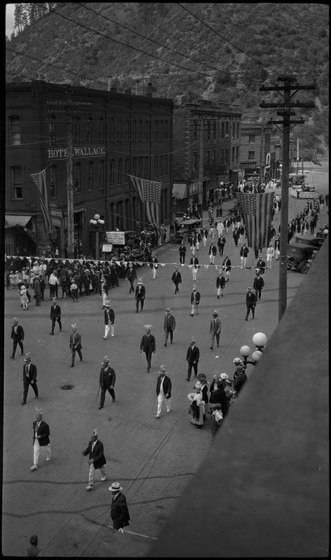 Men walking in the Benevolent and Protective Order of the Elks parade. They are wearing hats with fake antlers. Spectators watch from the side of the street. Garlands hang across the street.
