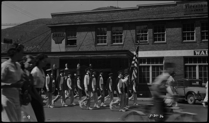 Men in uniform walking in Memorial Day parade. One of the men is holding an American flag. Three girls are walking by on the left. A boy rides by on his bicycle.
