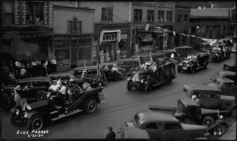 Series of cars in procession during the Benevolent and Protective Order of Elks parade. People are sitting in the cars and American flags are attached to the first two cars. Spectators are watching from the sidewalk.