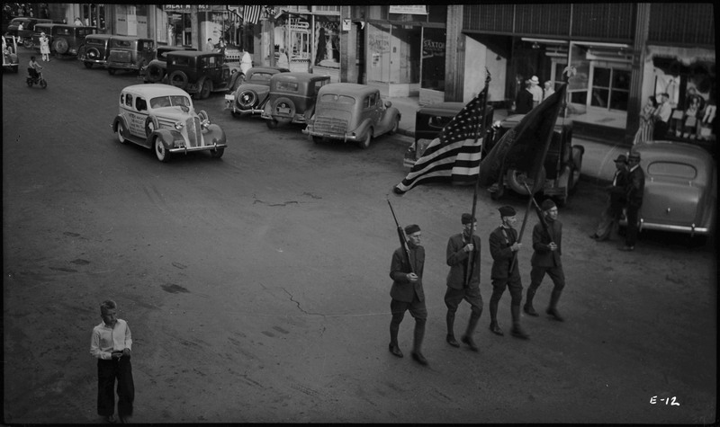 Four men, two holding rifles and the other two holding flags, in the Benevolent and Protective Order of Elks parade. A boy walks on the left. Spectators watch from the sidewalk.