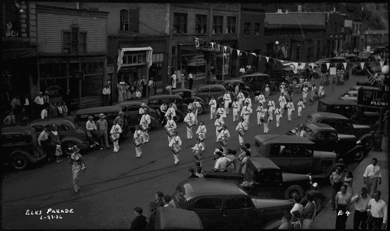 Children in a marching band during the Benevolent and Protective Order of Elks parade. They are wearing white uniforms and holding instruments. Spectators watch near parked cars along the street.