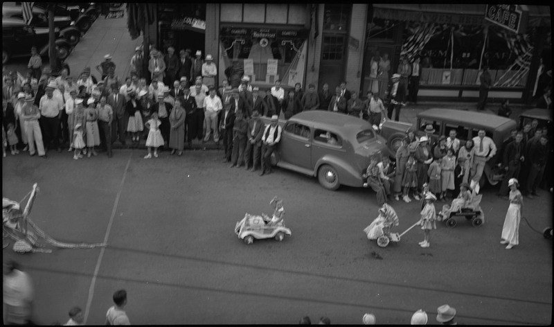 Children in a toy car, wagon, and stroller in the Benevolent and Protective Order of Elks parade. Spectators watch from the sidewalk and parked cars along the street.