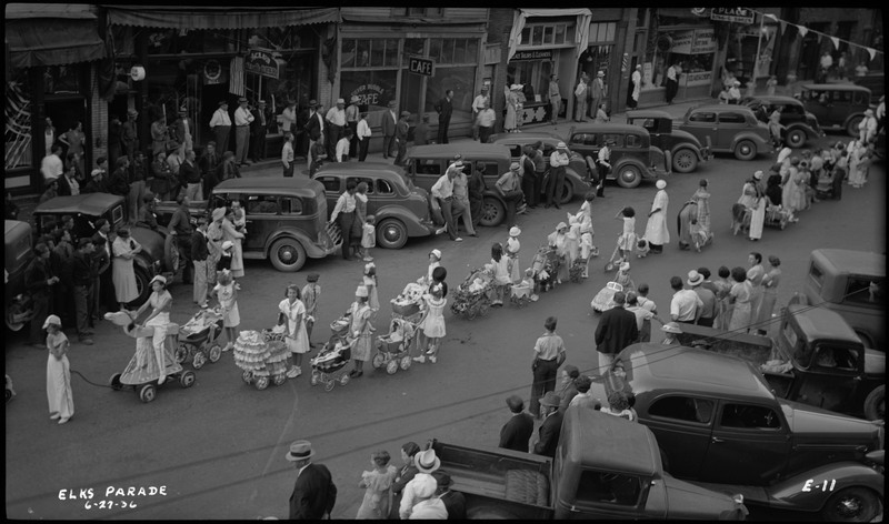 Children and several adults walking with strollers in Benevolent and Protective Order of Elks parade. Spectators watch from the sidewalk and parked cars along the street.