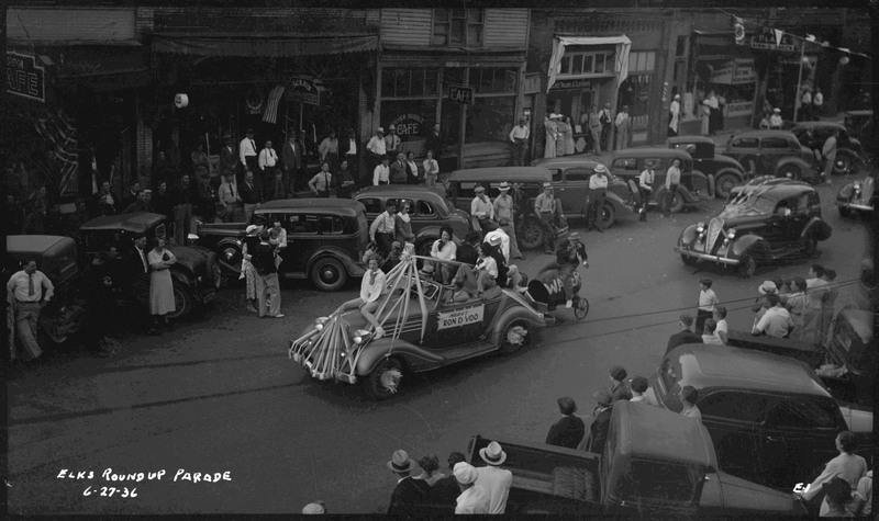 Several women and men in a small automobiles with the text, "Mark's Ron D Voo" on the side. A person is sitting on a water barrel is attached to the back of the vehicle. Other vehicles follow this first automobile. Spectators watch the Benevolent Protective and Order of Elks roundup parade from the sidewalk or parked cars along the side of the street.