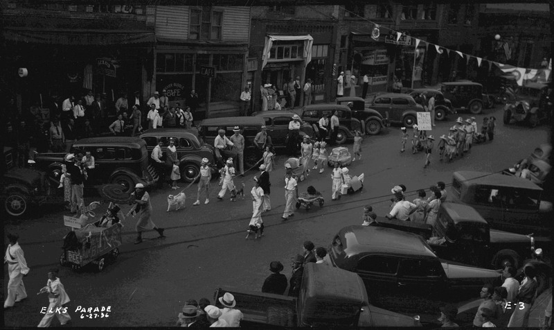 Children walking (some with strollers, two walk a dog) during the Benevolent and Protective Order of Elks parade. Spectators watch from the sidewalks and near parked cars along the street.