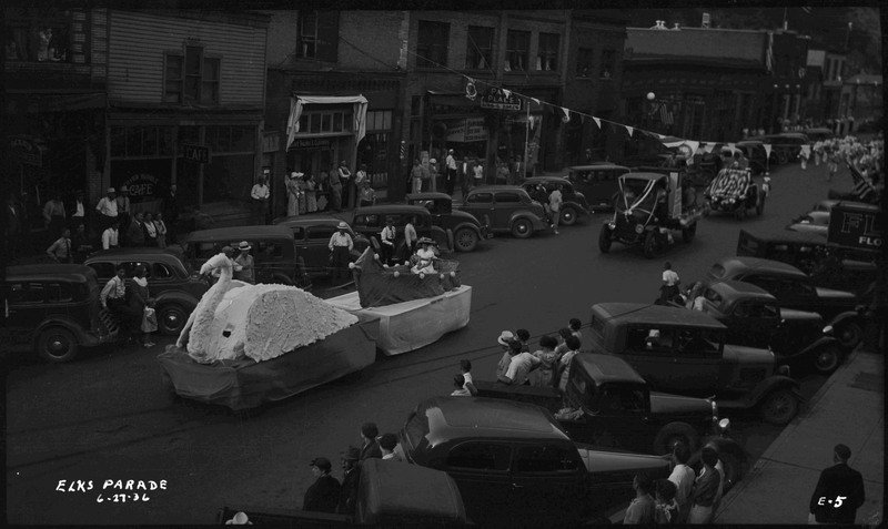 A woman sits in a fake boat on a float with a large swan during the Benevolent and Protective Order of Elks parade. Other floats and vehicles follow this swan float. Spectators watch from the sidewalk and near cars that are parked along the street.