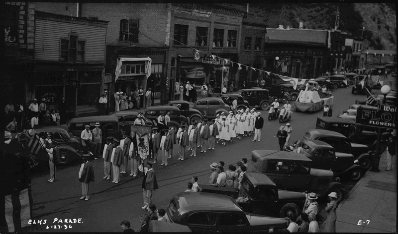Men in uniforms with the number 54 embroidered to their identical capes stand in the Benevolent and Protective Order of Elks parade. Spectators watch from the sidewalk and near parked cars. Two motorcyclists, swan float, and more vehicles follow these uniformed men. 