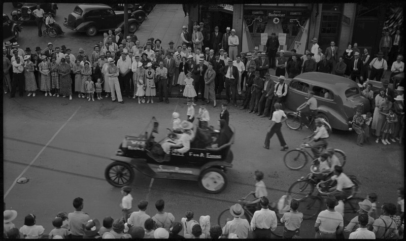 Several people sit in a vehicle as several boys follow on bicycles during the Benevolent and Protective Order and Elks parade. Spectators gather to watch from the sides.